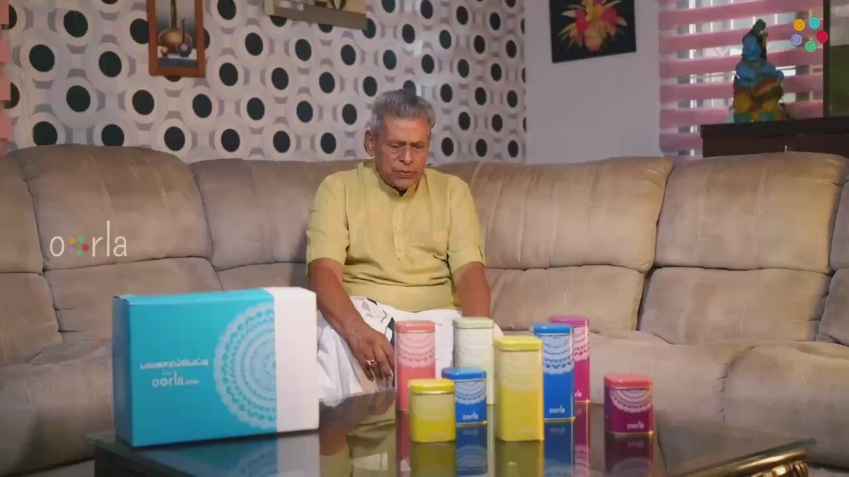 A still from the film: an elderly man in a yellow kurta sitting at home, talking across a table laid with an Oorla sweet box and a row of printed snack tins.